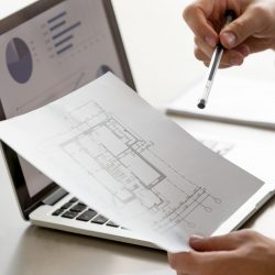 Close up view of male hand holding project plan, financial statistics data on laptop screen at background, analyzing real estate market, mortgage loan with low rate, construction investment