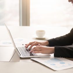 Close-up side view of serious businesswoman working at office desk on laptop, typing and looking at screen, surrounded by diagrams and calculating charts. Business, finances, economics and technology