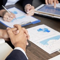 Crop shot of coworking businesspeople sitting at table with paper charts and tablet having meeting